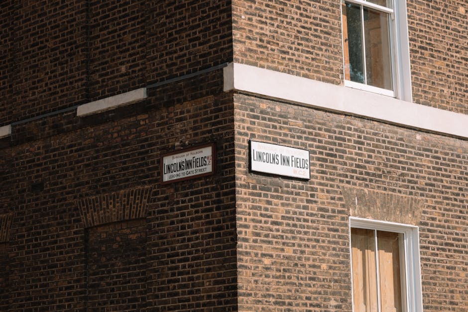 A brick building with a corner view, featuring two street name signs: one reading 'Lincoln's Inn Fields' and the other indicating the location leads to Gate Street. The building has a white horizontal band separating the upper and lower levels, and a window on the lower floor with a partly boarded-up covering. In the foreground, there is a loading area or pavement where a removal process may take place. Although no furniture, boxes, or removal equipment are visible in this image, the setting suggests a typical London property exterior that could be involved in home relocation or furniture transport activities, with natural daylight illuminating the scene. The presence of the signs indicates a well-known street, relevant to household removals and moving logistics with Man with Van Holborn often operating within this area, supporting packing and moving services.