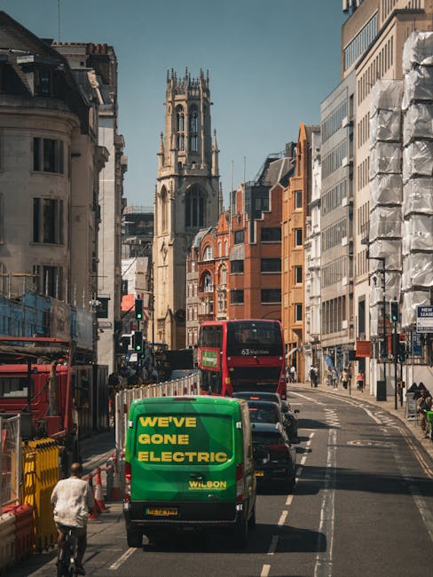 A busy urban street scene during daytime featuring a variety of vehicles including a green van with the text 'We've Gone Electric' and a red double-decker bus, with nearby cars and bicycles. The street is lined with multi-storey buildings, some under construction or renovation, and a prominent historic church tower visible in the background. The street appears to be part of a city centre, with people walking and vehicles moving through the area. This setting relates to house removals and moving services provided by Man with Van Holborn, highlighting the logistics involved in efficient property relocation and furniture transport within an urban environment.