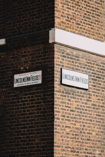 A brick building with a corner view, featuring two street name signs: one reading 'Lincoln's Inn Fields' and the other indicating the location leads to Gate Street. The building has a white horizontal band separating the upper and lower levels, and a window on the lower floor with a partly boarded-up covering. In the foreground, there is a loading area or pavement where a removal process may take place. Although no furniture, boxes, or removal equipment are visible in this image, the setting suggests a typical London property exterior that could be involved in home relocation or furniture transport activities, with natural daylight illuminating the scene. The presence of the signs indicates a well-known street, relevant to household removals and moving logistics with Man with Van Holborn often operating within this area, supporting packing and moving services.
