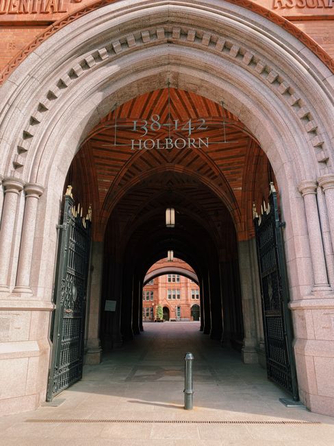 A view of an arched entrance to a historic building in Holborn, with the address number 1342 and the name 'Holborn' displayed above. The arch is constructed from stone with decorative columns on either side, supporting a curved brick ceiling with a wooden finish. Inside the archway, a narrow passageway extends into the distance, illuminated by hanging light fixtures. On either side of the entrance, there are black metal gates partially open, revealing an area where furniture, cardboard boxes, and packing materials are visible, indicating preparations for home relocation or moving services. A metal bollard is positioned on the pavement just outside the arch. The overall scene reflects a professional environment involved in the logistics of house removals, including loading or unloading activities for a moving company such as Man with Van Holborn.