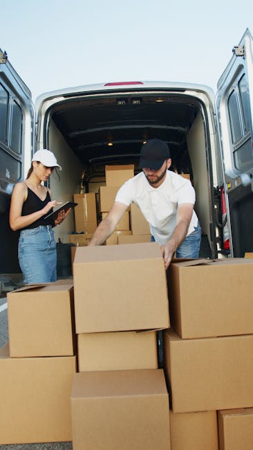 Two individuals are engaged in a home relocation or furniture transport process, unloading cardboard boxes from the open rear doors of a large white van, situated on a paved area possibly outside a property. The man, wearing a black cap, white shirt, and jeans, is actively handling a box, lifting or placing it onto a stack, while the woman, dressed in a white cap, black top, and jeans, holds a clipboard or tablet, likely for inventory or tracking purposes. Inside the van, additional stacked boxes are visible, organized for loading or unloading, with some protected by plastic wrap. The vehicle's interior is dark, contrasting with the bright outdoor environment, and the scene captures a moment in packing and moving logistics, with equipment such as trolleys or straps not visibly present. This setup aligns with professional removals services, emphasizing the careful handling and transportation of household items, as provided by Man with Van Holborn, supporting efficient and secure moving operations.