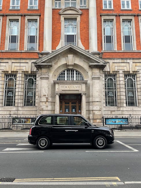 A black van parked on the street in front of a historic brick building with large sash windows and decorative stonework around the main entrance, which features a stone arch and Roman numeral inscript