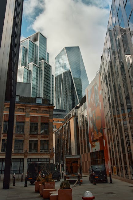 A city street scene featuring modern high-rise office buildings with glass facades reflecting the sky and surrounding structures, adjacent to a lower, older brick building. The narrow street is lined 