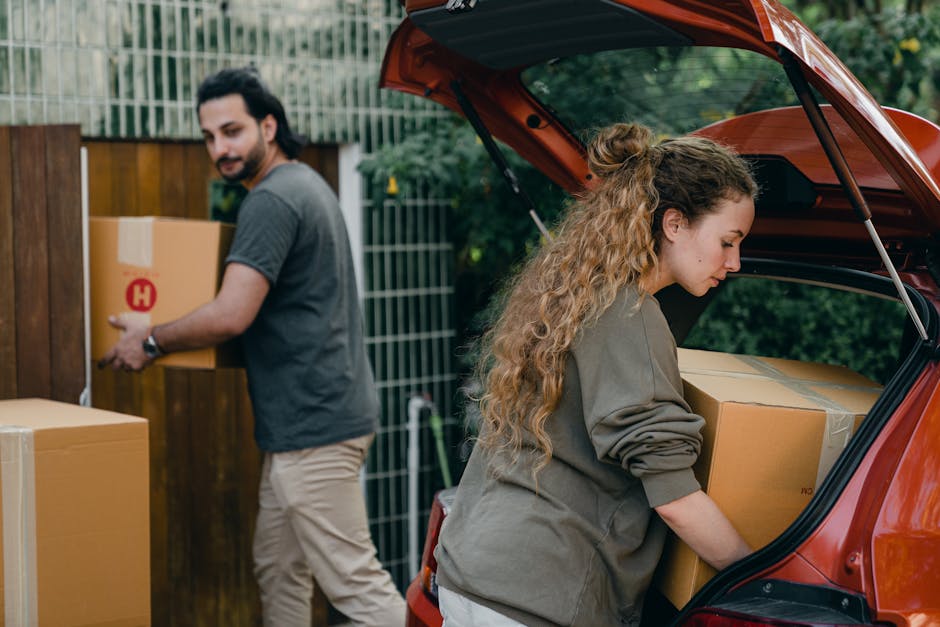 A woman with long, curly blonde hair wearing a grey long-sleeved top is seen loading a cardboard box into the open boot of a small, orange hatchback vehicle parked outside a property. Inside the vehicle, other boxes are visible, some wrapped in packing material. In the background, a man with dark hair, a beard, wearing a grey t-shirt and beige trousers, is walking towards the vehicle holding a similar cardboard box with a red 'H' label. The setting appears to be a residential area with a wooden fence and greenery behind, indicating a garden or backyard. The scene captures a home relocation or furniture transport process involving packing and loading boxes, supported by the presence of packing materials, furniture, and the vehicle used for moving. Man with Van Holborn offers services related to removals, assisting with efficient moving logistics and packing.