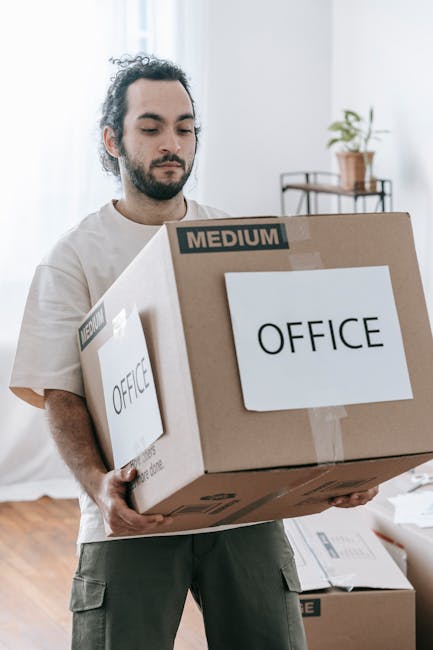 A man with a beard and tied-back dark hair is inside a well-lit room, holding a large cardboard box labeled 'OFFICE' with medium weight indicated on the top. The box appears to contain office furniture or supplies and is wrapped with brown packing tape. In the background, there are additional moving boxes, some lying on the wooden floor and others stacked, with packaging labels visible. The room has light-colored walls, and a small metal shelving unit with a potted plant is positioned near a window that allows natural daylight to brighten the space. This scene captures the packing and loading process involved in office relocation services offered by [COMPANY_NAME], which specialises in professional removals and furniture transport, supporting smooth business moves and minimising downtime during office moves in Holborn.