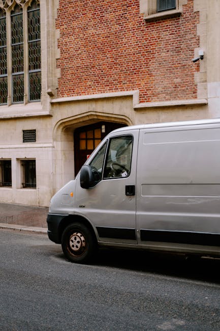 A silver delivery van parked on a city street in front of a historic brick building with large stained glass windows and stone architectural details. The van is positioned close to the sidewalk, with its side door closed, indicating a possible relocation or furniture transport process associated with home moves or packing activities. The surrounding environment includes a paved road with a curb, and the building's entrance features an arched doorway with a small window above it. The lighting appears natural, suggesting daytime. This scene reflects the typical setting for loading or unloading household items during house removals, with the van prepared for transporting furniture and boxes as part of the moving logistics handled by Man with Van Holborn.