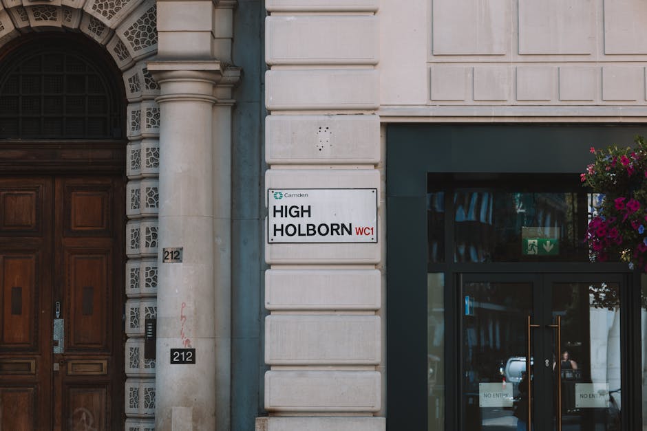 Close-up of the exterior corner of a building showing a white sign reading 'HIGH HOLBORN WC1' mounted on a beige stone wall, adjacent to a large wooden door with intricate carvings and a black keypad lock. To the right, there is a glass entrance door with a black frame, featuring a small green emergency exit sign and a floral arrangement with pink and purple flowers hanging above it. The area is well-lit with natural daylight, highlighting the building’s architectural details such as ornamental stonework around the door and textured stone blocks on the wall. The scene suggests a typical urban property entrance, relevant to house removals, with the presence of a moving crew potentially preparing for a furniture transport or home relocation from this location. The signage indicates this is a designated address segment within Holborn, supporting services like packing, loading, and transportation involved in professional removals by [COMPANY_NAME].